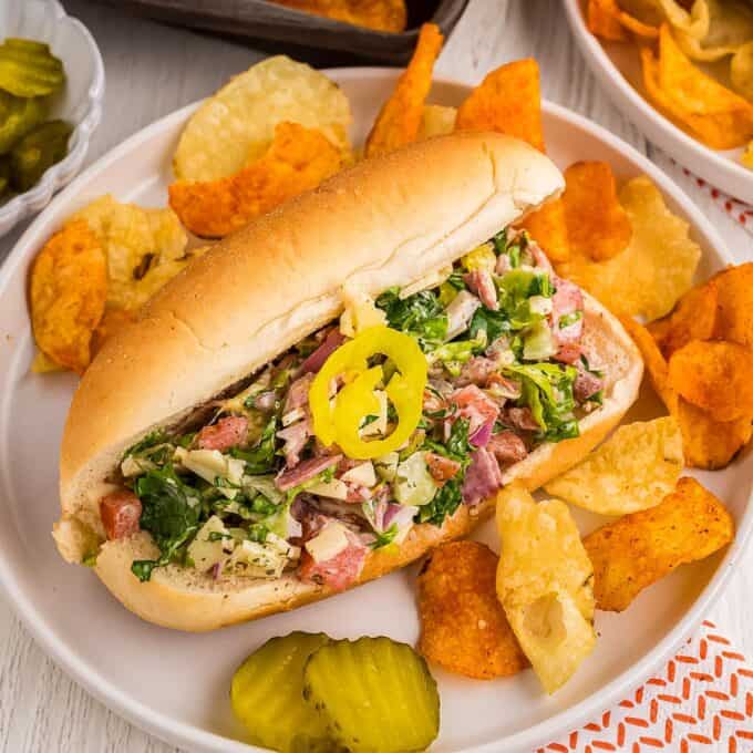 chopped salad sandwich on a white plate with chips and pickles.