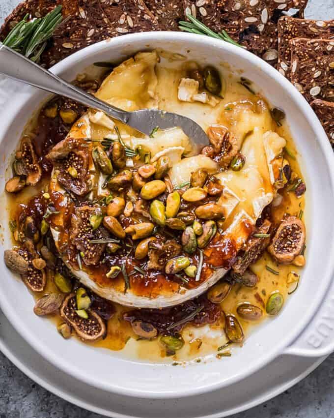 This honey herb and fig baked brie recipe is perfect for the holidays, or any party! A wheel of brie is topped with sweet fig jam, fresh rosemary, dried fig slices, salty pistachios, and sticky honey, then baked until perfectly gooey. Serve with baguette slices, apples, crackers, and more!