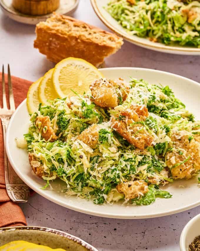 two brussels sprouts caesar salads on white plates with crusty bread.