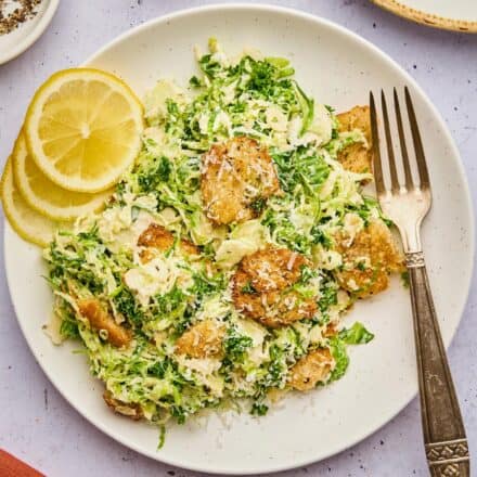 brussels sprouts caesar salad on a white plate with a fork and lemon slices.